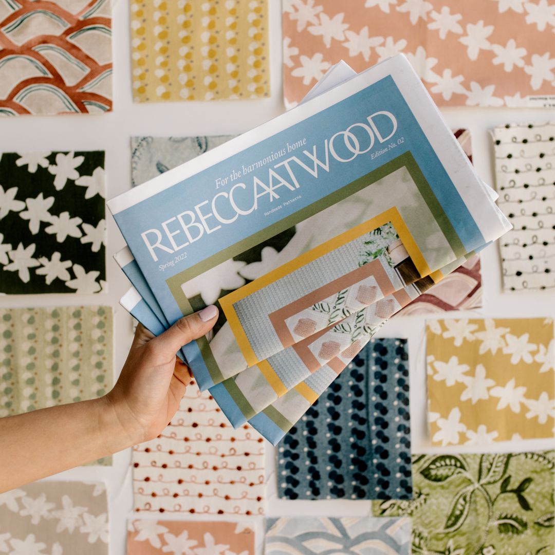 An extended arm holding copies of a newspaper with colouful patterns on the front page. Text on the front page reads: "For the harmonious home - Rebecca Atwood"